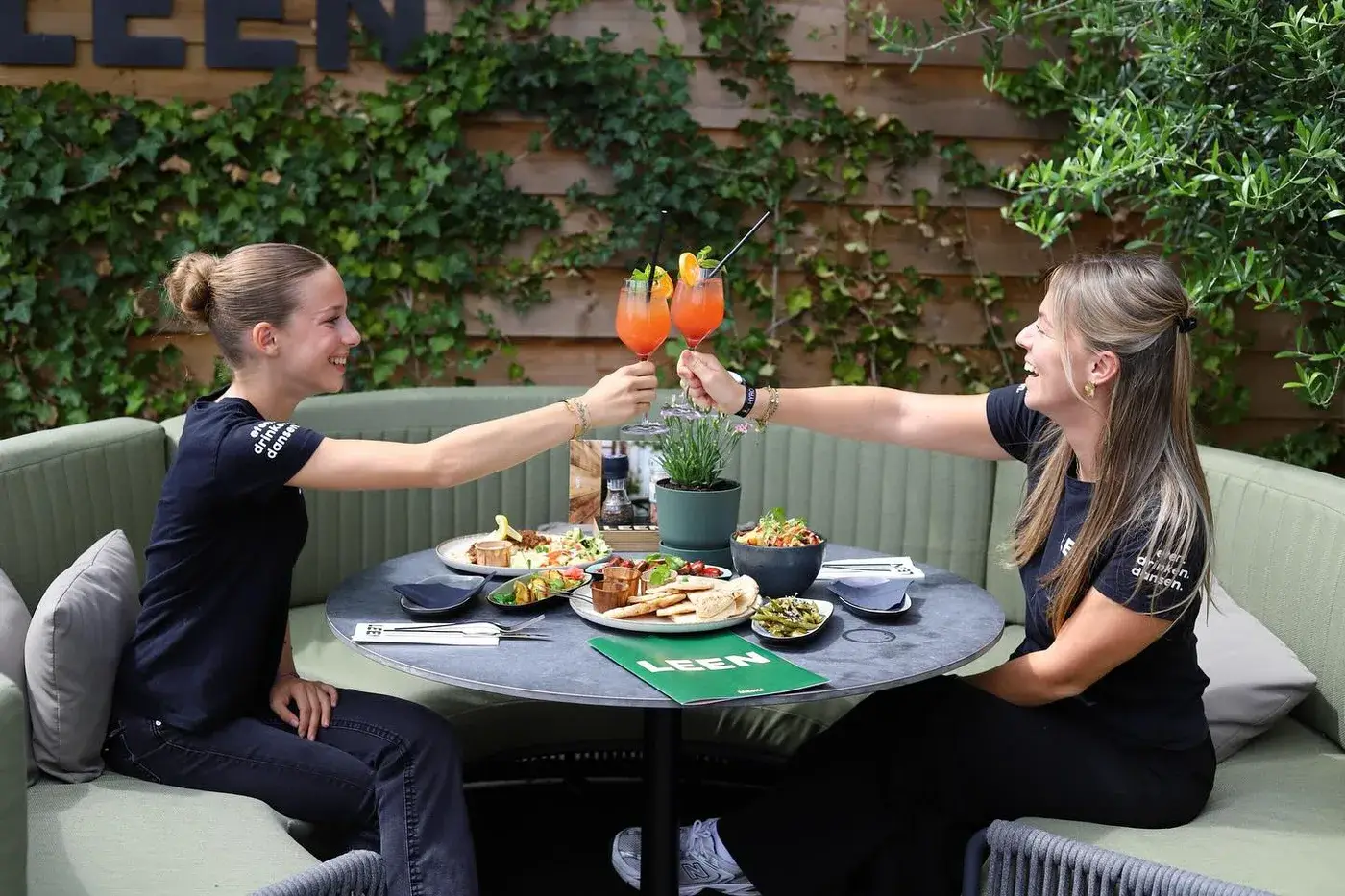 Twee gasten proosten met cocktails in de loungebank op het terras van Club LEEN